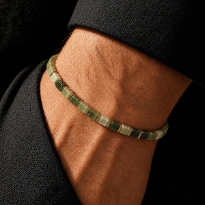 May include: A bracelet composed of small, square-shaped beads in varying shades of sage and forest green. The beads are arranged in a continuous line, separated by thin, metallic-toned spacers, creating a uniform and structured appearance.