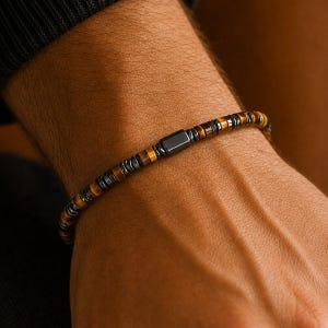 May include: A beaded bracelet featuring alternating small, flat, cylindrical tiger eye beads in shades of brown and metallic gray hematite beads, centered with a single, smooth, rectangular black bead as a focal point.