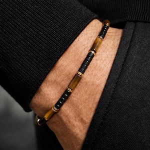 Könnte beinhalten: Ein Perlenarmband mit einem sich wiederholenden Muster aus kleinen, zylindrischen schwarzen Perlen und längeren, glatten braunen Tigeraugen-Steinen. Die Perlen sind durch dünne, goldfarbene Metall-Abstandshalter getrennt, was ein strukturiertes Design ergibt.