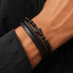 Op de afbeelding: Een set van drie zwarte armbanden bestaande uit een gladde leren band, een gevlochten leren band en een kralenarmband met een kleine zwarte kruishanger. De kralen zijn rond en matzwart en het kruis heeft een getextureerd oppervlak.