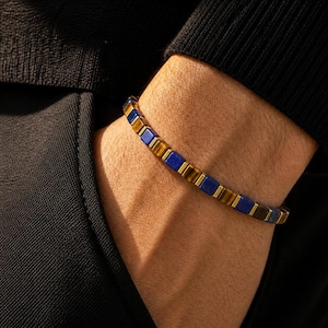 Puede incluir: Una pulsera de cuentas con cuentas cuadradas alternas en piedra ojo de tigre azul profundo y marrón dorado, dispuestas en un patrón repetitivo sobre un cordón flexible, creando una apariencia estructurada y geométrica alrededor de la muñeca.
