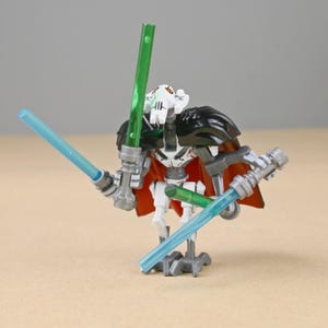 May include: A General Grievous action figure with a white and black head, red cape, and black shoulder armor. The figure is holding four lightsabers, two green and two blue.