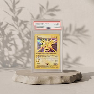 May include: A 1999 Pokemon Game Zapdos holographic trading card encased in a clear protective slab. The card features a yellow background, 90 HP, and the attacks Thunder and Thunderbolt. The label at the top reads 1999 POKEMON GAME ZAPDOS HOLO #16 GEM MT.