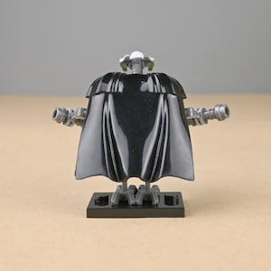 May include: A black Lego minifigure of General Grievous, featuring a black cape, gray arms with blasters, and a detailed head. The figure stands on a black base, showcasing a Star Wars theme.