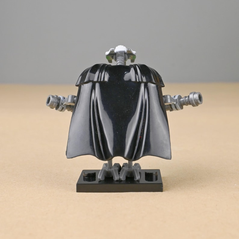 May include: A black Lego minifigure of General Grievous, featuring a black cape, gray arms with blasters, and a detailed head. The figure stands on a black base, showcasing a Star Wars theme.