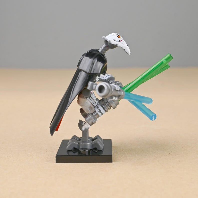 May include: A General Grievous action figure, featuring a white head with orange markings, a black cape, and silver armor. The figure is holding two lightsabers, one green and one blue, and is mounted on a black base.