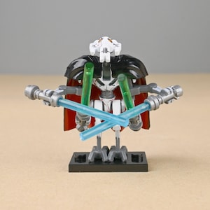 May include: A General Grievous action figure, featuring a white head with black accents, a black cape, and a red cloak. The figure holds four lightsabers, two green and two blue, and is mounted on a black base.