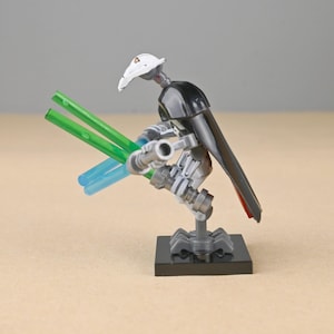 May include: A General Grievous action figure, featuring a white helmet, black cape, and multiple arms holding green and blue lightsabers. The figure is posed on a black base, showcasing a detailed design.