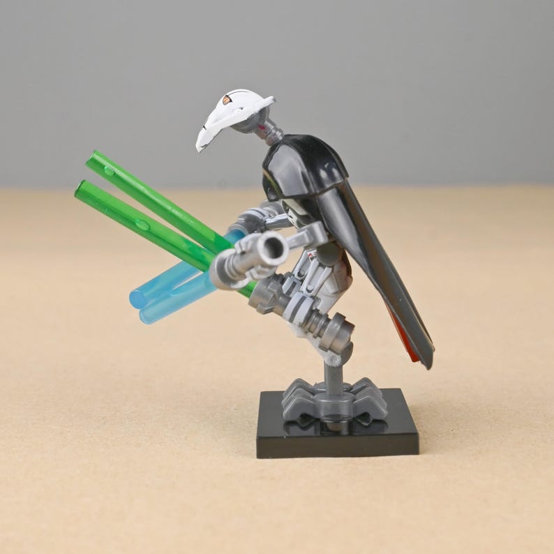 May include: A General Grievous action figure, featuring a white helmet, black cape, and multiple arms holding green and blue lightsabers. The figure is posed on a black base, showcasing a detailed design.
