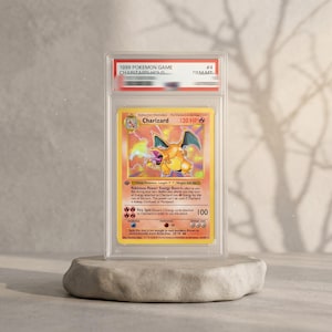 May include: A 1999 Pokemon Game Charizard holographic trading card encased in a clear protective slab. The card features the orange dragon-like creature, 120 HP, and the text Charizard, Fire Spin, and Energy Burn. The slab label reads GEM MT.