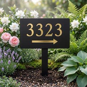 May include: A black address sign with gold numbers and an arrow. The numbers "3323" are prominently displayed. The arrow points to the right. The sign is mounted on a black post and set in a garden with flowers.