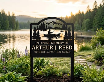 May include: A black metal memorial garden stake featuring a silhouette of a person fishing in a boat on a lake with mountains and pine trees. The text reads IN LOVING MEMORY OF ARTHUR J. REED OCTOBER 12, 1947 - MAY 4, 2023 in white lettering.