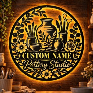 May include: A circular black metal wall sign with warm backlighting featuring a pottery studio theme. The design includes a large vase, bowls, and sculpting tools surrounded by floral motifs. The text reads CUSTOM NAME Pottery Studio.