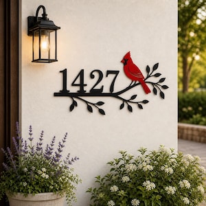 Puede incluir: Letrero de dirección de metal negro con el número 1427, un pájaro cardenal rojo y ramas con hojas. El letrero está montado en una pared blanca junto a una lámpara exterior negra y plantas en macetas.