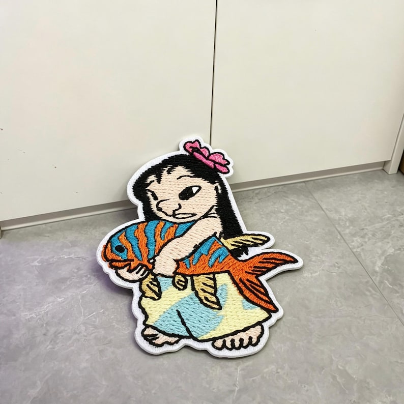 May include: An embroidered patch featuring a cartoon character with black hair and a pink flower, wearing a light yellow dress with blue patterns, holding a large orange fish with blue stripes. The design has a thick white border outline.
