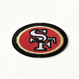 Op de afbeelding: Een ovaalvormige geborduurde patch met de letters SF in het wit met een zwarte omlijning. De letters zijn gecentreerd op een rode achtergrond, omringd door een dunne gouden rand en een dikke zwarte buitenrand, wat een embleem vormt.