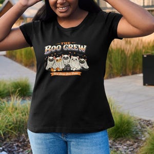 Op de afbeelding: Een zwart t-shirt met korte mouwen en een afbeelding van vijf katten verkleed als spoken. De tekst THE BOO CREW staat boven de katten en Nine Lives, Zero Fears op een oranje banner eronder, inclusief kleine sterretjes en vleermuizen.