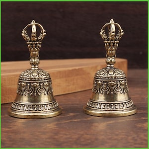Peut inclure: Deux cloches antiques de couleur bronze avec des d&eacute;tails orn&eacute;s. Chaque cloche pr&eacute;sente une poign&eacute;e en forme de couronne et des motifs complexes autour du corps de la cloche. Les cloches sont expos&eacute;es sur une surface en bois.