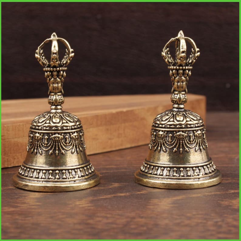 Peut inclure: Deux cloches antiques de couleur bronze avec des d&eacute;tails orn&eacute;s. Chaque cloche pr&eacute;sente une poign&eacute;e en forme de couronne et des motifs complexes autour du corps de la cloche. Les cloches sont expos&eacute;es sur une surface en bois.