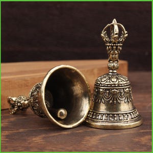 Peut inclure: Deux cloches antiques en bronze avec des d&eacute;tails orn&eacute;s. L'une est debout, avec une poign&eacute;e d&eacute;taill&eacute;e et des motifs complexes. L'autre est couch&eacute;e, r&eacute;v&eacute;lant le battant. Les cloches ont une esth&eacute;tique vintage.