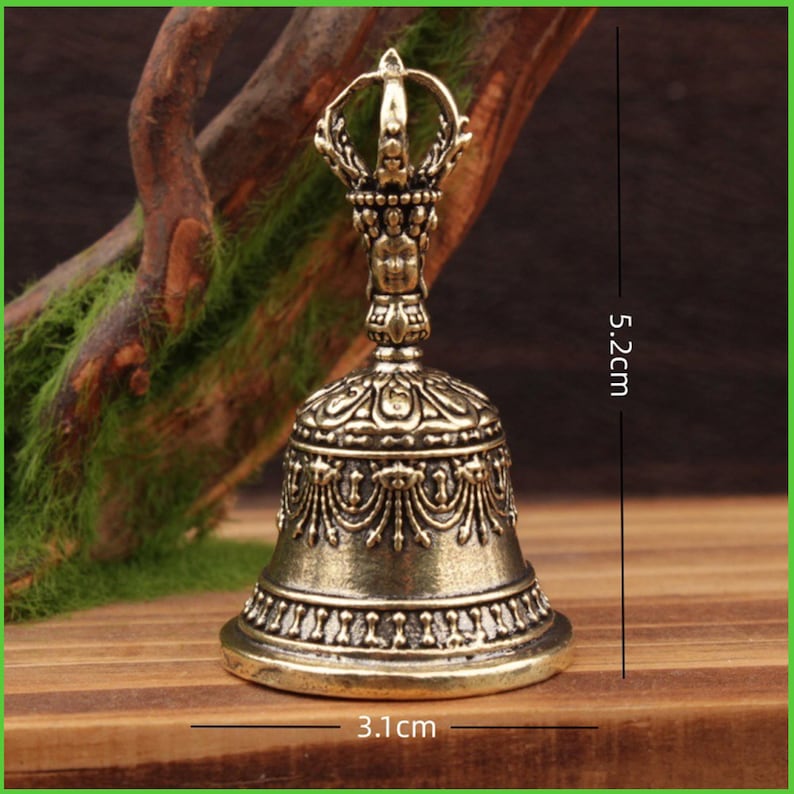 Peut inclure: Une cloche antique en laiton, de 5,2 cm de haut et 3,1 cm de large, avec des motifs en relief complexes. La cloche pr&eacute;sente une poign&eacute;e d&eacute;taill&eacute;e avec un motif en forme de couronne. La cloche est expos&eacute;e sur une surface en bois avec un fond naturel.