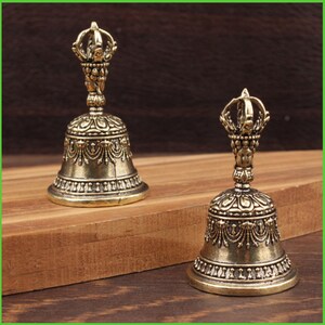 Peut inclure: Deux cloches antiques de couleur bronze avec des d&eacute;tails orn&eacute;s. Chaque cloche pr&eacute;sente une poign&eacute;e d&eacute;corative avec un motif en forme de couronne. Les cloches sont plac&eacute;es sur une surface en bois, mettant en valeur leurs motifs complexes et leur aspect vieilli.