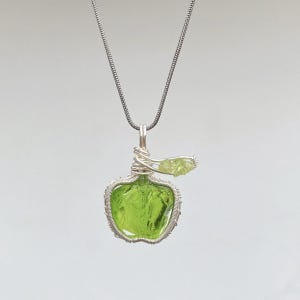 May include: A silver wire-wrapped pendant necklace featuring a green peridot gemstone in the shape of an apple. The pendant hangs from a silver chain. The apple-shaped gemstone is a vibrant, light green color.