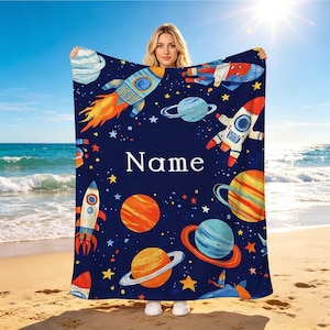 Puede incluir: Una manta rectangular de color azul oscuro con un colorido estampado de temática espacial que incluye varios planetas, estrellas y cohetes. La palabra Name está impresa en letras mayúsculas blancas sin remates en el centro de la manta.