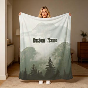 Puede incluir: Una manta suave con un paisaje impreso de montañas verdes brumosas y pinos oscuros en la base. El centro de la manta muestra el texto Custom Name en una fuente oscura, negrita y sin remates sobre un fondo claro y con niebla.