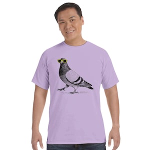 Pode incluir: Uma t-shirt de manga curta em violeta claro com uma ilustração gráfica a preto e branco de um pombo a usar óculos de sol amarelo-brilhante. A ave está representada de perfil, de pé sobre uma linha de solo texturada no peito.