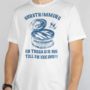 May include: A white short-sleeved t-shirt featuring a blue graphic of an open can of surströmming. The text reads SURSTRÖMMING at the top and EN TUGGA GÖR DIG TILL EN VIKING!!! at the bottom, with additional small text on the can label.
