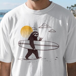 Peut inclure: T-shirt blanc avec un paresseux de dessin animé portant une planche de surf. Le paresseux est brun foncé, avec un soleil jaune et des nuages en arrière-plan. Le paresseux fait le signe shaka.