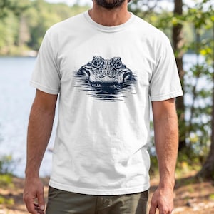 Può includere: T-shirt bianco con un'illustrazione blu scuro della testa di un alligatore che emerge dall'acqua. Il disegno dettagliato è centrato sulla maglietta, creando un contrasto deciso. Adatto per abbigliamento casual.