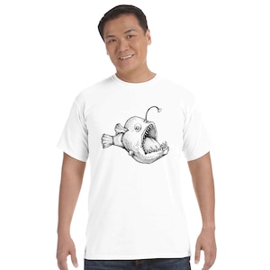 Op de afbeelding: Een wit T-shirt met korte mouwen en een zwart-witte illustratie in inktstijl van een zeeduivel. De afbeelding toont de vis met een grote open mond, scherpe tanden en een opvallend lokaas dat uit de kop van de vis steekt.