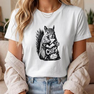 Könnte beinhalten: Weißes T-Shirt mit einem schwarz-weißen Motiv eines Eichhörnchens, das eine Chipstüte hält. Auf der Tüte steht "CHIPS" und "0% AUTHORITY". Das T-Shirt ist aus einem weichen Material.