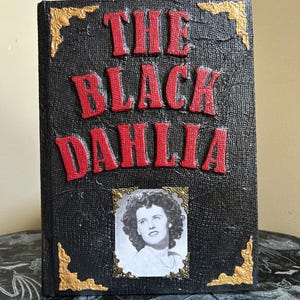 K&ouml;nnte beinhalten: Ein schwarzes Buch mit dem Titel "THE BLACK DAHLIA" in roten, erhabenen Buchstaben. Ein Schwarz-Wei&szlig;-Foto einer Frau ist in Gold gerahmt. Goldene Eckakzente zieren den Einband des Buches.