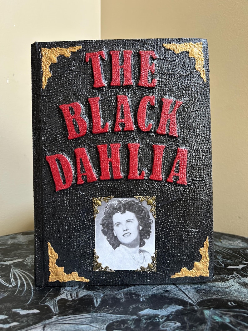 K&ouml;nnte beinhalten: Ein schwarzes Buch mit dem Titel "THE BLACK DAHLIA" in roten, erhabenen Buchstaben. Ein Schwarz-Wei&szlig;-Foto einer Frau ist in Gold gerahmt. Goldene Eckakzente zieren den Einband des Buches.