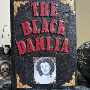 K&ouml;nnte beinhalten: Ein dekoratives Buch mit dem Titel "THE BLACK DAHLIA" in roter, erhabener Schrift. Das Buch hat einen schwarzen, strukturierten Einband mit goldenen Eckakzenten und ein gerahmtes Schwarz-Wei&szlig;-Foto einer Frau.