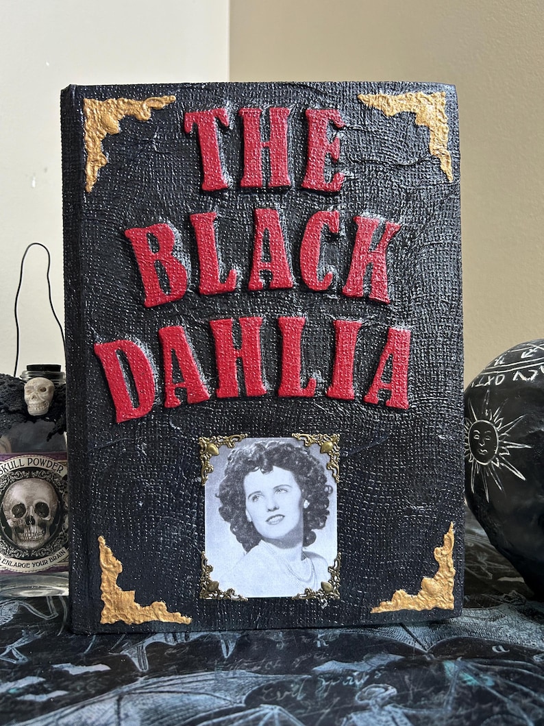 K&ouml;nnte beinhalten: Ein dekoratives Buch mit dem Titel "THE BLACK DAHLIA" in roter, erhabener Schrift. Das Buch hat einen schwarzen, strukturierten Einband mit goldenen Eckakzenten und ein gerahmtes Schwarz-Wei&szlig;-Foto einer Frau.