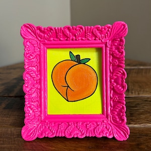 May include: A small framed painting of a peach with a green stem and leaf. The peach is painted in a cartoon style with a bright orange color. The frame is a bright pink color.