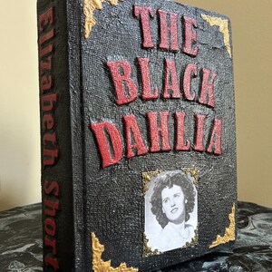 K&ouml;nnte beinhalten: Dekoratives Buch mit dem Titel "The Black Dahlia" in roten, erhabenen Buchstaben auf einer strukturierten schwarzen Oberfl&auml;che. Das Buch zeigt ein Schwarz-Wei&szlig;-Portr&auml;t und goldene Eckakzente. Der Name des Autors, Elizabeth Short, ist vertikal auf dem Buchr&uuml;cken aufgedruckt.