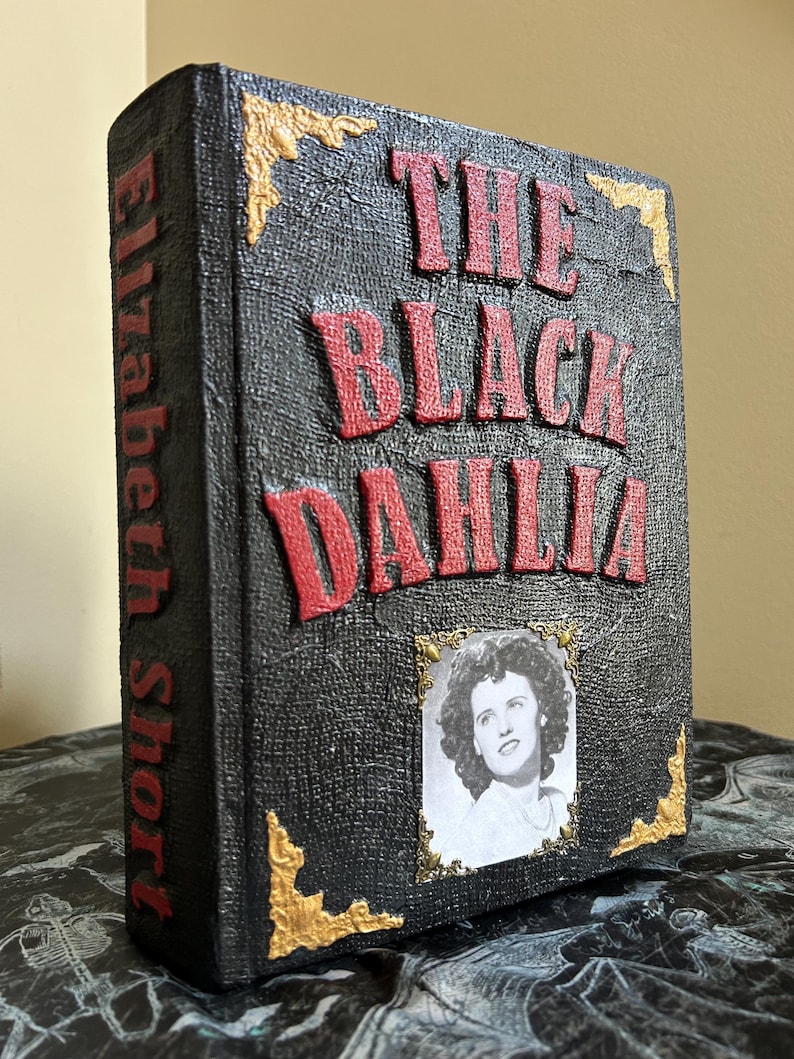 K&ouml;nnte beinhalten: Dekoratives Buch mit dem Titel "The Black Dahlia" in roten, erhabenen Buchstaben auf einer strukturierten schwarzen Oberfl&auml;che. Das Buch zeigt ein Schwarz-Wei&szlig;-Portr&auml;t und goldene Eckakzente. Der Name des Autors, Elizabeth Short, ist vertikal auf dem Buchr&uuml;cken aufgedruckt.