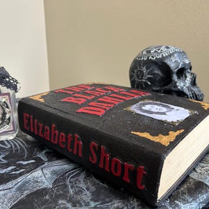 K&ouml;nnte beinhalten: Ein schwarzes Buch mit dem Titel "The Black Dahlia" von Elizabeth Short, mit roten Buchstaben und goldenen Akzenten. Ein sch&auml;delartiges Glas mit der Aufschrift "Skull Powder" und eine schwarze Totenkopf-Figur sind ebenfalls zu sehen, was ein gruseliges, gotisches Thema erzeugt.