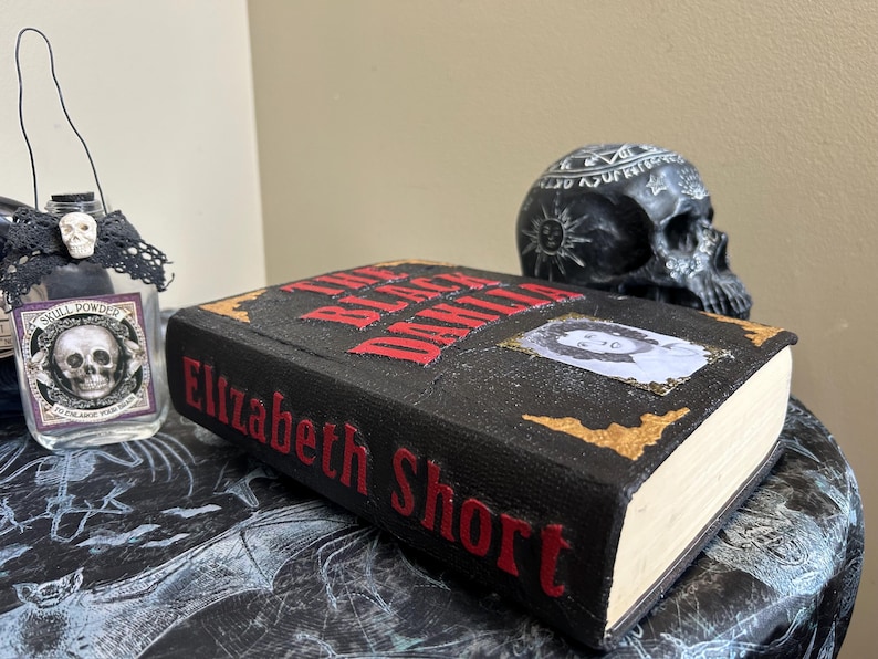 K&ouml;nnte beinhalten: Ein schwarzes Buch mit dem Titel "The Black Dahlia" von Elizabeth Short, mit roten Buchstaben und goldenen Akzenten. Ein sch&auml;delartiges Glas mit der Aufschrift "Skull Powder" und eine schwarze Totenkopf-Figur sind ebenfalls zu sehen, was ein gruseliges, gotisches Thema erzeugt.