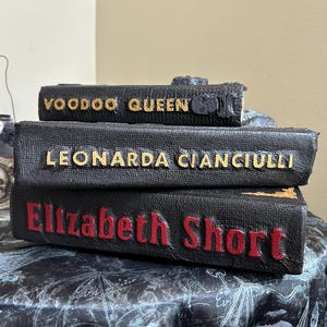 K&ouml;nnte beinhalten: Drei gestapelte B&uuml;cher aus schwarzem Kunstleder mit goldenen und roten Buchstaben. Das oberste Buch tr&auml;gt die Aufschrift "Voodoo Queen", das mittlere Buch "Leonarda Cianciulli" und das untere Buch "Elizabeth Short". Die B&uuml;cher haben eine strukturierte Oberfl&auml;che.