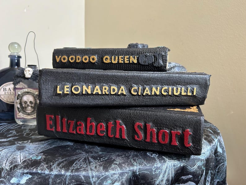 K&ouml;nnte beinhalten: Drei gestapelte B&uuml;cher aus schwarzem Kunstleder mit goldenen und roten Buchstaben. Das oberste Buch tr&auml;gt die Aufschrift "Voodoo Queen", das mittlere Buch "Leonarda Cianciulli" und das untere Buch "Elizabeth Short". Die B&uuml;cher haben eine strukturierte Oberfl&auml;che.
