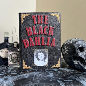 K&ouml;nnte beinhalten: Ein Stillleben mit einem Buch mit dem Titel "The Black Dahlia" mit roter Schrift, einem Foto und goldenen Akzenten. Ein schwarzer Sch&auml;del und Apothekerflaschen vervollst&auml;ndigen das Arrangement.