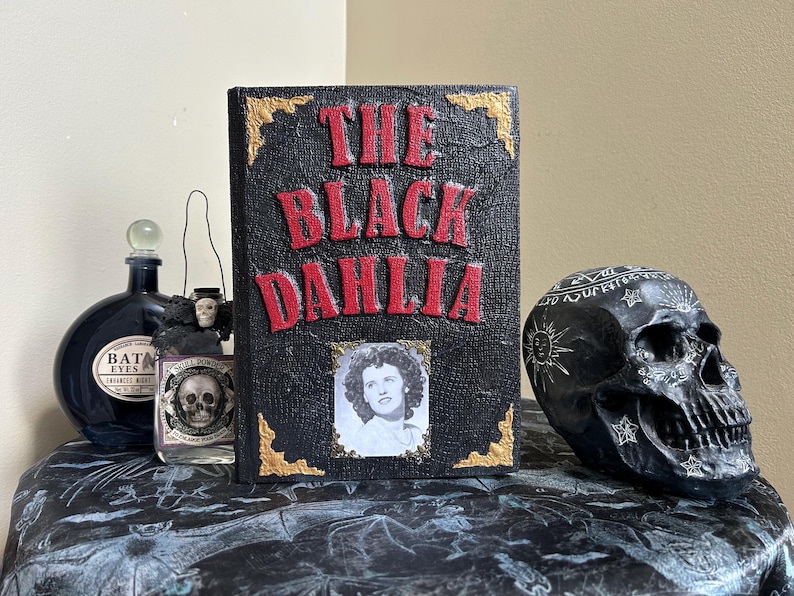 K&ouml;nnte beinhalten: Ein Stillleben mit einem Buch mit dem Titel "The Black Dahlia" mit roter Schrift, einem Foto und goldenen Akzenten. Ein schwarzer Sch&auml;del und Apothekerflaschen vervollst&auml;ndigen das Arrangement.