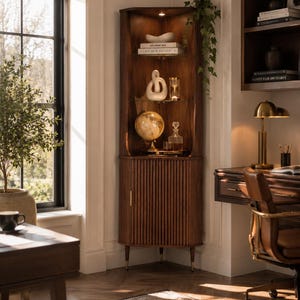 May include: A dark wood corner cabinet with a fluted door and brass-tipped legs. The cabinet displays decorative items like a globe, books, and a white sculpture. A desk and chair are visible in the background, creating a home office setting.