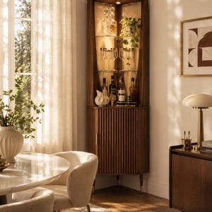 May include: A wooden corner cabinet with glass shelves, displaying glassware, bottles, and a small plant. The cabinet has a fluted door and is illuminated from within. The room also features a round table, chairs, and a side cabinet.